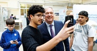 Frontale Aufnahme einer Person und einer jugendlichen Person, die nebeneinander stehen. Die jugendliche Person zeigt der anderen Person etwas auf ihrem Handy. Dahinter sind verschiedene Schilder einer Messe