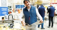 Frontale Aufnahme einer Person, die neben einem Tisch steht, ein dünnes Metallbrett hält und mit einer Metallschere in das Metall schneidet