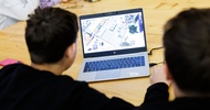 Rückenansicht zweier Personen, die nebeneinander an einem Holztisch sitzen und gemeinsam auf den Bildschirm eines Laptops blicken. Auf dem Bildschirm ist ein Warenlager