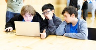 Drei Kinder sitzen nebeneinander an einem Holztisch und blicken auf den Bildschirm eines Laptops. Daneben steht eine Person. Im Hintergrund sind weitere Personen