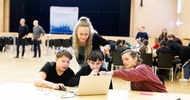 Zwei Kinder sitzen nebeneinander an einem Holztisch und blicken auf den Bildschirm eines Laptops. Neben ihnen sitzt eine Person, die auf den Laptop zeigt. Daneben steht eine weitere Person. Im Hintergrund sind weitere Personen