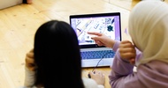 Rückenansicht zweier Personen, die nebeneinander an einem Holztisch sitzen und gemeinsam auf den Bildschirm eines Laptops blicken. Auf dem Bildschirm ist ein Warenlager