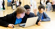 rei Kinder sitzen nebeneinander an einem Holztisch und blicken auf den Bildschirm eines Laptops. Im Hintergrund sind weitere Personen