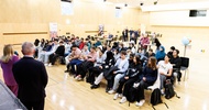 In einem großen Saal mit Holzboden und Holzvertäfelungen an der Wand sitzen in einem Publikum viele Jugendliche. Ihnen gegenüber stehen zwei Personen in Sakkos. Die linke Person spricht in ein Mikrofon. Auf einem Stelltisch im Hintergrund liegt ein Globus