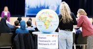 Auf einem Stelltisch liegt ein Globus. Am Stelltisch ist ein Zettel angeklebt, auf dem Station Teamwork- und Netzwerk-Profi steht. Im Hintergrund ist die Rückenansicht eines sitzenden Publikums. Dem Publikum gegenüber steht eine Person