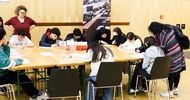 In einem großen Raum mit Holzboden sitzen um einen großen Tisch viele jugendliche Personen, die jeweils Dinge auf Zetteln notieren. Neben ihnen stehen zwei erwachsene Personen. Auf einem Banner neben ihnen steht Ihre Cargo, unsere Lösungen