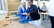 Eine erwachsene und eine jugendliche Person stehen nebeneinander an einem Tisch in einer Werkstatt. Auf dem Tisch liegen verschiedene Werkzeuge. Die erwachsene Person erklärt etwas