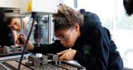 Ein Kind mit Schutzbrille bedient den Hebel eines Metallbohrers. In der Bohrmaschine ist ein kleines Objekt eingespannt