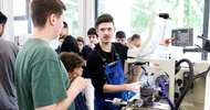 Eine Person mit Schutzbrille und blauer Latzhose steht neben einem Gerät in einer Werkstatt und bedient den kleinen Display des Geräts. Neben ihr stehen ein Kind und eine jugendliche Person, die zu der anderen Person blicken. Im Hintergrund sind weitere Personen