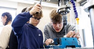 Ein Kind mit Schutzbrille bedient den Hebel eines Metallbohrers. Neben ihr steht eine weitere Person, die ein Objekt unter dem Bohrer mit einer Hand festhält und darauf blickt