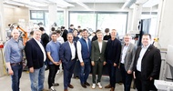 Porträtaufnahme von neun Personen in einer Werkstatt, die dicht nebeneinander stehen. Im Hintergrund sind verschiedene Geräte sowie mehrere Personen, die im Raum stehen