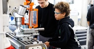 Ein Kind mit Schutzbrille bedient ein Gerät in einer Werkstatt mit einer Kurbel, die an der Seite des Geräts befestigt ist. Neben ihr steht eine weitere Person