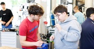  Zwei jugendliche Personen stehen nebeneinander an einem Tisch in einer Werkstatt. Auf dem Tisch ist ein Werkzeug montiert