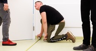 Eine Person hält das Ende eines Maßbands an die untere Wand eines Raumes. Daneben stehen zwei weitere Personen
