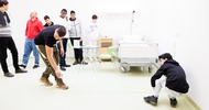 In einem Raum mit einem Krankenhausbett stehen mehrere Personen. Zwei Personen messen mit Maßband eine Fläche am Boden