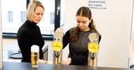 Eine Person mit geschlossenen, dunklen Haaren steht neben einem Zapfhahn. Neben ihr steht eine Person mit kinnlangen, blonden Haaren, die ihr hilft. Auf einem Tresen vor ihnen steht ein Glas Bier