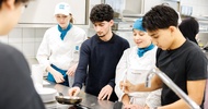 In einer Gastronomieküche stehen an einem Ceranfeld zwei jugendliche Personen sowie zwei Personen in Kochkleidung. Die jugendlichen Personen haben jeweils vor sich eine Pfanne