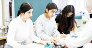 Drei Teenager stehen nebeneinander an einem Tisch. Vor ihnen sind verschiedene Stoffe. Ein Teenager malt mit einem roten Stift. Ihnen gegenüber steht eine erwachsene Person, die etwas erklärt