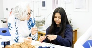 Ein Teenager sitzt an einem Tisch. Auf dem Tisch sind verschiedene Stoffe und Wollknäuel. Direkt neben dem Teenager ist eine Person mit weißen Haaren und Brille, die sich nach vorne beugt und etwas erklärt