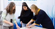 An einem Tisch stehen zwei erwachsene Personen und ein Teenager. Eine der erwachsenen Personen schneidet mit einem Messer ein Muster auf einem Blatt Papier aus. Die anderen beiden sehen zu