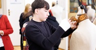 Ein Teenager in einem dunklen Pullover steht in einem Raum neben einer Modepuppe und bindet dieser ein Tuch um den Hals. Im Hintergrund sind weitere Personen
