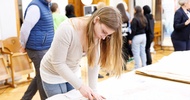 Ein Teenager mit Brille und offenen, hellen Haaren steht an einem Tisch und schneidet mit einem kleinen Messer ein Muster aus einem Blatt Papier aus. Im Hintergrund stehen weitere Teenager sowie erwachsene Personen