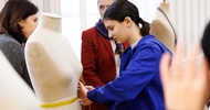 Ein Teenager misst mit einem Maßband die Taille einer Modepuppe. Neben ihr stehen zwei erwachsene Personen sowie eine weitere Modepuppe
