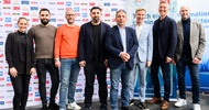 Porträt von acht Personen, die dicht nebeneinander vor großen Werbeplakaten stehen