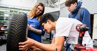 In einer Werkstatt steht ein Autoreifen auf einem Tisch. Der Reifen wird von einer Person in einer Latzhose festgehalten. Ein Teenager beugt sich nach vorne, blickt auf den Reifen und hat seine Hände auf dem Reifen. Daneben steht ein weiterer Teenager