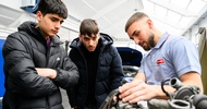 In einer Werkstatt stehen neben einem Motor eine erwachsene Person und zwei Teenager, die alle auf den Motor blicken. Im Hintergrund steht ein Auto mit geöffneter Motorhaube