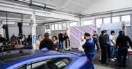 In einer Werkstatt parkt ein blaues Auto. Neben dem Auto stehen mehrere Personen und Jugendliche in kleinen Gruppen