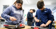 Ein Teenager steht an einem Tisch und schlägt mit einem Hammer einen breiten Nagel in eine MEtallplatte. Daneben steht ein weiterer Teenager, der einen Nagel hält