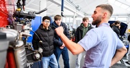 In einer Werkstatt steht eine Person neben einem Motor eines Fahrzeugs und redet. Der Person gegenüber stehen drei Teenager, die zuhören. Im Hintergrund steht ein Auto mit geöffneter Motorhaube