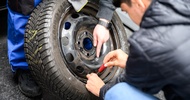 Nahaufnahme des Reifens eines Autos, bei dem eine Person eine Felge festschraubt. Die Hand einer anderen Person deutet auf die Felge