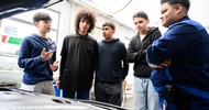Neben einer geöffneten Motorhaube eines Fahrzeugs stehen eine erwachsene Person und vier Jugendliche, die sich unterhalten