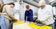 An einem Tisch in einer Werkstatt stehen zwei Personen, die auf Autoglasscheiben Saugknöpfe halten. Daneben steht eine Person in einem blauen Overall