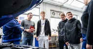 Neben einer geöffneten Motorhaube eines Fahrzeugs stehen drei erwachsene Personen und drei Teenager, die reden, in einer Werkstatt