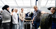 In einer Werkstatt steht ein Autoreifen auf einem Tisch und wird von einer erwachsenen Person mit der Hand festgehalten. Der Person gegenüber sind zwei Teenager und zwei erwachsene Personen