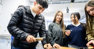 Ein Teenager steht an einem Tisch und schlägt mit einem Hammer einen breiten Nagel in eine MEtallplatte. Daneben stehen drei weitere Teenager