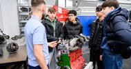 Ein Motor eines Fahrzeugs steht in einer Werkstatt. Um den Motor stehen vier Teenager und eine erwachsene Person. Die Teenager blicken zur erwachsenen Person