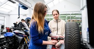  In einer Werkstatt steht ein Autoreifen erhöht. Eine Person fixiert eine Felge in der Mitte des Reifens. Neben der Person steht ein Teenager. Im Hintergrund stehen Motorräder