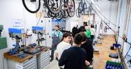 In einem Raum mit Drechslermaschinen stehen viele Jugendliche neben einer Holztischplatte. Neben einer Drechslermaschine steht eine Person in Arbeitskleidung