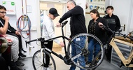 In einem Raum hebt eine Person ein schwarzes Fahrrad in die Höhe. Direkt daneben steht eine jugendliche Person. Neben den beiden stehen weitere Jugendliche und eine Person, die zu ihnen blicken