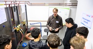In einem Raum verlaufen an einer Wand verschiedene Rohre. Im Raum steht eine Gruppe Jugendlicher. Ihnen gegenüber ist eine weitere Person, die spricht. An der Wand hängt ein Zettel, auf dem Kälteanlagentechnik steht