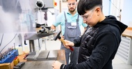 Seitliche Aufnahme einer jugendlichen Person mit Schutzbrille, die neben einer Tischplatte steht und einen Hammer in der rechten Hand hält