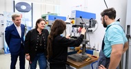 Eine Person mit langen Haaren steht neben einer Drechslermaschine. Direkt neben ihr steht eine weitere Person mit Schutzbrille. Links neben den beiden stehen zwei weitere Personen, die zusehen