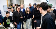Person mit Schutzbrille steht neben einem Gerät. Ihr gegenüber stehen viele weitere Personen. Die Person mit der Schutzbrille redet zu den anderen Personen