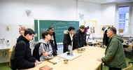 Hinter einem Tisch stehen sechs Jugendliche. Ihnen gegenüber steht eine Person, die etwas erklärt. An einer Tafel im Hintergrund steht Sattlerei