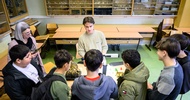 Hinter einem Tisch steht eine Person, die etwas erklärt. Dem Tisch gegenüber stehen Personen
