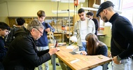 Eine jugendliche Person sägt mit einer Handsäge an einem Stück Holz. Andere Personen und Jugendliche stehen neben ihr und schauen zu